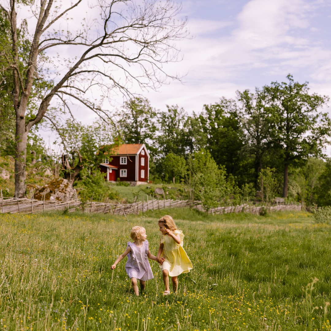 Två barn springer över en sommaräng i Stensjö by, med ett rött hus i bakgrunden